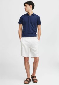 Marine polo skjorte laget av bomull, hvite linshorts og brune sandaler med stropper. Modellen står med hendene i lomma, og vinkler kroppen litt til venstre.