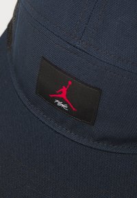 Marineblå kasket lavet af tekstureret stof, med et sort mærke med et rødt "Jumpman" logo og hvid tekst, syede detaljer, flad skygge.