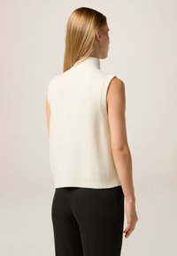 Suéter sin mangas de cuello alto en color crema con dobladillo acanalado. Textura suave, forma ajustada. Combinado con pantalones negros, mostrando elementos de diseño minimalistas.