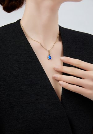 Collier en chaîne dorée avec un pendentif en forme de larme orné d'une pierre bleue entourée de petites pierres claires, porté par une personne vêtue d'un haut noir à col en V.