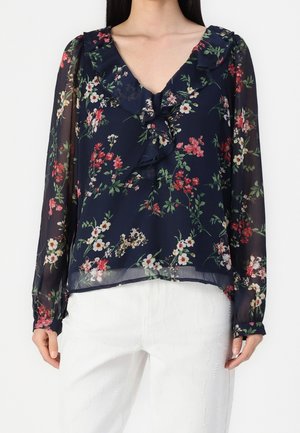 Granatowa przezroczysta bluzka z różowo-białym kwiatowym wzorem, falbaniastym dekoltem w kształcie litery V i długimi rękawami, noszona z białymi spodniami.