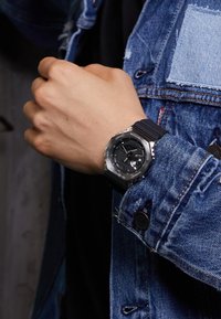 Montre numérique noire avec un cadran rond, une lunette en métal et un bracelet en caoutchouc, portée sur une main avec une veste en denim comportant des accents usés.