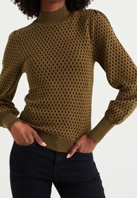 Kvinna i en olivgrön och svart prickig stickad tröja med puffärmar, ribbade mudd- och fållar, med händerna i fickorna på svarta byxor.