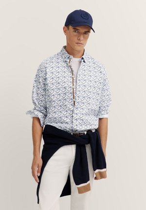 Floral-patterned shirt in white and blue, with a button-down collar and rolled sleeves. Navy cap with logo, paired with light-colored pants and a sweater.