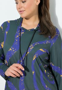 Kapuzenshirt aus dehnbarem, glattem Stoff mit einer dunkelgrünen Basis und wirbelnden Mustern in Blau und Gelb, umrandet mit schwarzen Kordeln.