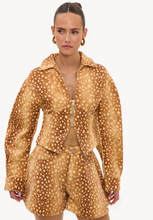 Femme aux cheveux attachés porte une veste marron à fermeture éclair et un short assorti, tous deux avec un motif blanc tacheté ressemblant à une fourrure d’animal.