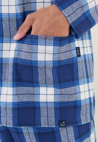 Camisa a cuadros azul y blanca con un bolsillo visible. Hecha de un tejido suave; presenta tonos variables de azul y una pequeña etiqueta de marca.