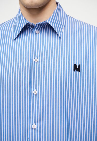 Homme portant une chemise boutonnée à rayures verticales bleues et blanches avec un col pointu et un "M" noir brodé sur la poitrine.