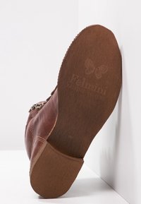 Hnědá kožená bota s zakřiveným tvarem, s kovovými akcenty a texturovanou gumovou podrážkou. Logo je embosováno na spodní straně.