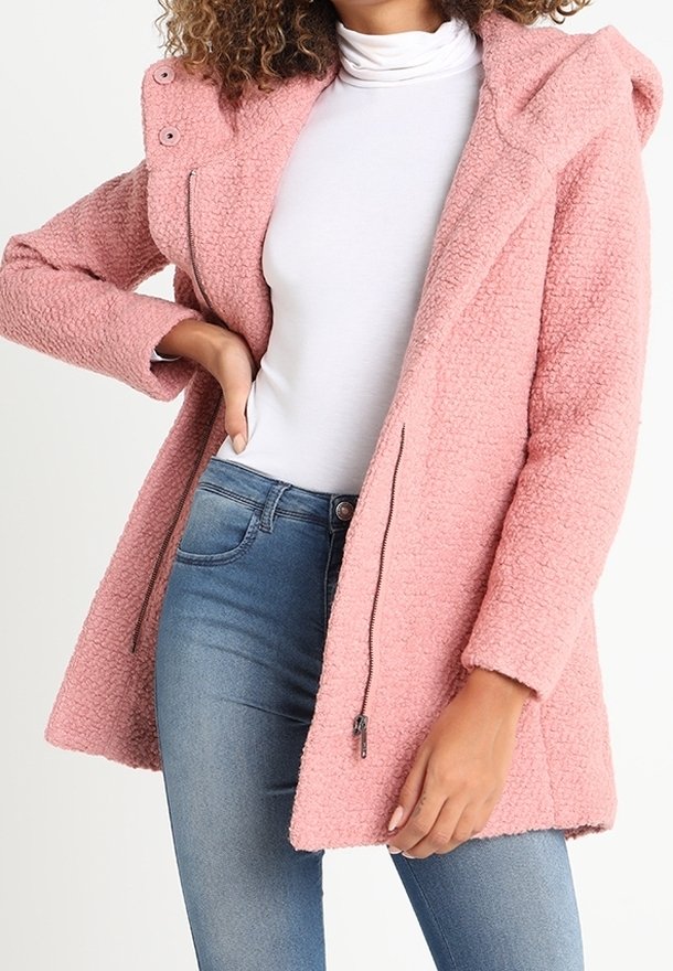 Femme portant un manteau à capuche rose texturé sur un col roulé blanc et un jean bleu, une main sur la hanche et l'autre détendue.