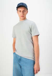Giovane uomo con un cappello blu, una maglietta a maniche corte grigia chiara e un jeans azzurro, con una mano in tasca su uno sfondo bianco.