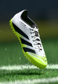 Botte de football blanche et noire avec des accents verts, présentant des rainures sur la tige et une semelle texturée avec des crampons proéminents, flottant au-dessus de l'herbe.