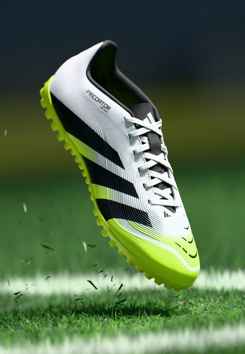Botte de football blanche et noire avec des accents verts, présentant des rainures sur la tige et une semelle texturée avec des crampons proéminents, flottant au-dessus de l'herbe.