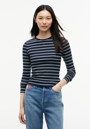 Jeune femme aux cheveux noirs et raides porte un t-shirt à manches longues rayé bleu marine et blanc et un jean bleu, une main dans la poche.