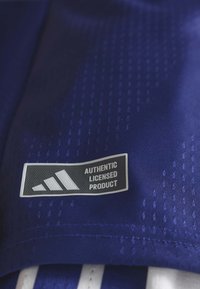 Modrá tkanina s hladkou textúrou má čierno-bielu nálepku s textom "AUTENTICKÝ LICENCOVANÝ PRODUKT" po boku loga Adidas.