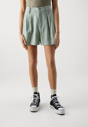 Light green linen shorts with pleats and a button closure. Paired with black high-top sneakers and beige socks. Minimalist design.