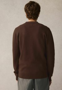 Pull tricot marron avec col rond et ourlet côtelé, associé à un pantalon gris à rayures fines, vu de dos. Texture lisse, design décontracté.