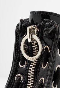 Gros plan sur une bottine en cuir verni noir avec une fermeture éclair en métal argenté et une tirette ronde gravée DKNY, lacets noirs passant à travers des œillets argentés.