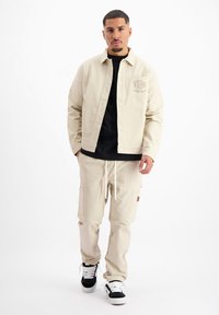 Beige katoenen jack met knoopsluiting en een borstzak, gecombineerd met bijpassende cargo-broek met zijzakken en een trekkoord. Zwarte sneakers.
