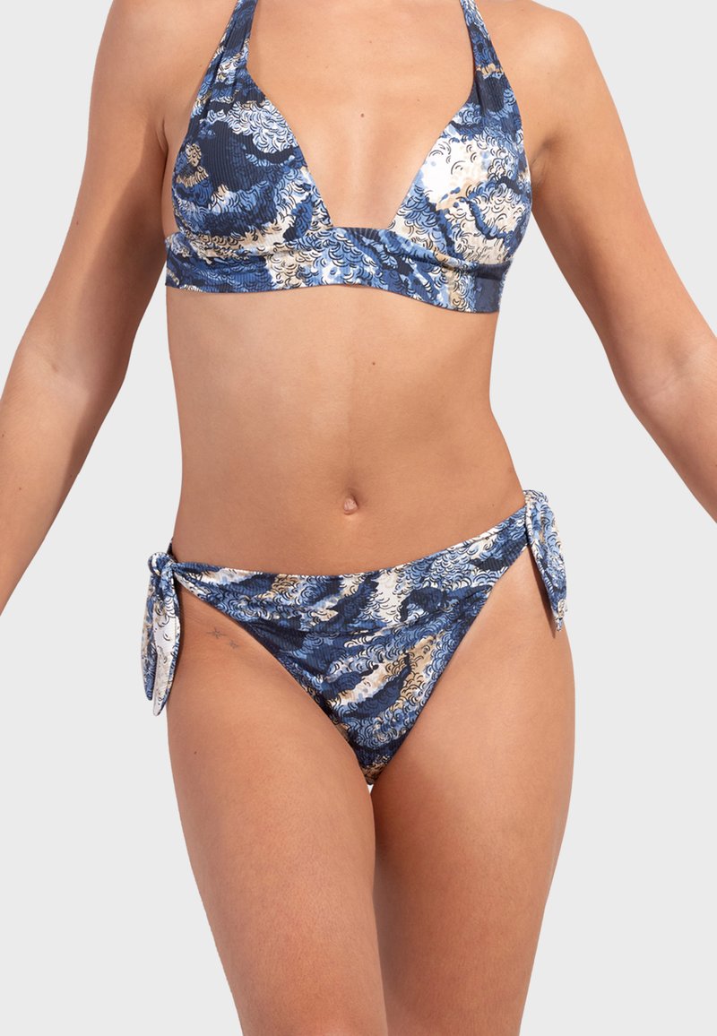 Femme portant un bikini bleu et blanc à motifs, avec un haut dos-nu et un bas avec nouettes sur les côtés, sur un fond clair uni.