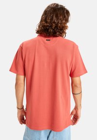 Kurzärmliges, korallenfarbiges Shirt aus weichem Stoff. Hat einen runden Halsausschnitt und ein kleines Markenetikett auf der Rückseite. Klassische Passform.