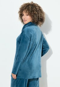 Top de terciopelo de manga larga en color verde azulado, con un cuello alto y un ajuste relajado, que presenta una textura suave y lisa.
