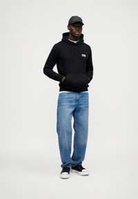 Μαύρη μα hoodie με μπροστινή τσέπη και κορδόνια, συνδυασμένη με ανοιχτό μπλε jeans σε χαλαρή γραμμή και μαύρα αθλητικά παπούτσια με λευκή σόλα.