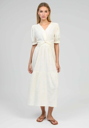 Femme debout dans une robe midi crème avec des manches bouffantes, un détail nœud à la taille et des sandales plates marron sur un fond gris uni.