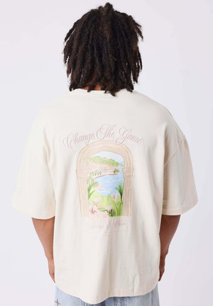 Personne aux cheveux bouclés foncés porte un T-shirt beige oversize avec une arche scénique colorée et le texte "Change The Game" dans le dos.