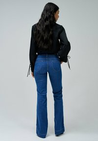 Blusa negra texturizada con mangas abullonadas y detalles de lazo, combinada con jeans de denim azul acampanados con bolsillos traseros y diseño de cintura alta.