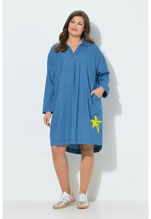 Frau trägt ein locker sitzendes blaues Denimkleid mit langen Ärmeln, einem gelben Seestern-Design auf der Tasche und silbernen Turnschuhen, steht vor weißem Hintergrund.