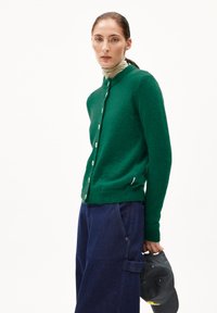 Vrouw in een groene knopen cardigan over een beige coltrui, donkere blauwe wijde broek, met een zwarte baseballpet tegen een witte achtergrond.