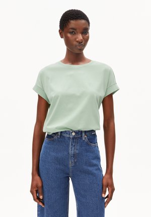 Hellgrünes Baumwoll-T-Shirt mit aufgerollten Ärmeln, rundem Ausschnitt und kleinem Logo, kombiniert mit hoch taillierter blauer Denim-Jeans, einfarbiger Hintergrund.