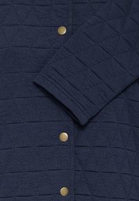 Tissu matelassé bleu marine avec des coutures géométriques, comprenant une manche et trois boutons-pression ronds en laiton sur une veste ou un manteau.