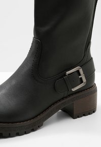 Botte noire montant jusqu'au genou en cuir synthétique avec une finition texturée. Dotée d'une boucle en métal et d'un talon en bois avec une semelle robuste.