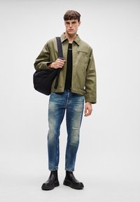 Olivengrønn jakke, svart t-skjorte, slitte blå jeans og svarte støvler. Modellen bærer en svart kviltet skulderveske.