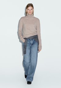 Maglione beige a collo alto con maniche lunghe, abbinato a jeans blu larghi e una camicia grigia legata in vita; modella che indossa scarpe nere.