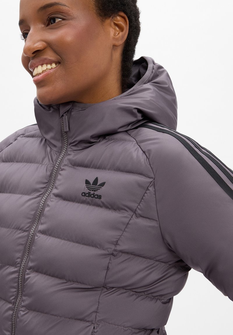 Persona sonriente vestida con una chaqueta de plumas gris de Adidas con capucha y franjas negras en las mangas, mirando ligeramente hacia la izquierda sobre un fondo blanco.