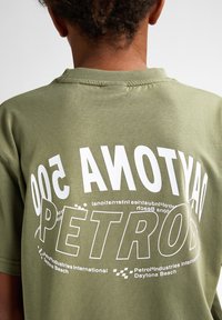 Olivově zelené tričko s krátkým rukávem se bílými grafikami s textem "DAYTONA 500 PETROS" a dalším textem. Jemná, bavlněná textura.