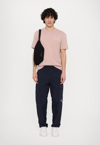 Lys rosa t-skjorte med korte ermer, marineblå cargobukser med to sidelommer, svart messengerbag og hvite joggesko. Glatt stofftekstur.
