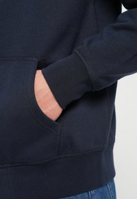 Námornícky modrý sveter s klokankou, rebrovanými manžetami a švami. Vyrobený z mäkkého, štruktúrovaného materiálu, s uvoľneným strihom.