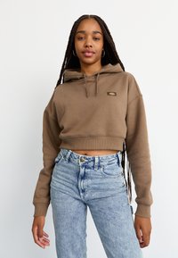 Hoodie corto marrone con tasca a marsupio, polsini a coste e una piccola etichetta con logo. Abbinato a jeans chiari a vita alta.