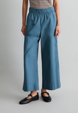 Osoba nosząca luźne niebieskie spodnie o szerokich nogawkach z elastycznym pasem oraz czarne baleriny typu Mary Jane, stojąca na jednolitym szarym tle.