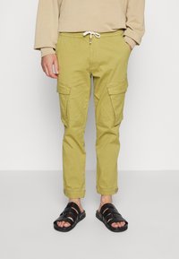 Calças cargo verde-oliva claras com bolsos laterais, punhos elásticos e cintura com cordão; usadas com sandálias pretas de tiras.