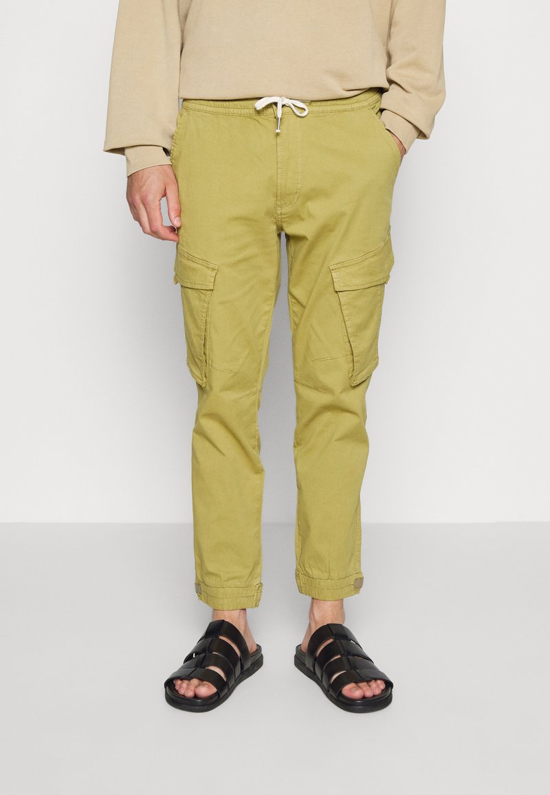 Calças cargo verde-oliva claras com bolsos laterais, punhos elásticos e cintura com cordão; usadas com sandálias pretas de tiras.