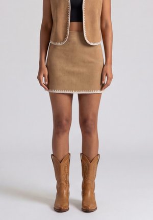 Donna con minigonna in camoscio beige e gilet senza maniche abbinato, top corto nero e stivali da cowboy in pelle beige, in piedi su sfondo grigio.