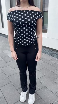 Juodas off-shoulder detaljeruotas marškinėlis su baltomis taškeliais, derintas su juodomis kelnėmis, turinčiomis auksinius akcentus, ir baltais sportbačiais.