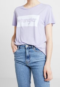 Fialové bavlněné tričko s krátkými rukávy a bílým obdélníkovým grafickým prvkem, na kterém je tučným písmem napsáno "Wrangler". V kombinaci se světle modrými džínami.