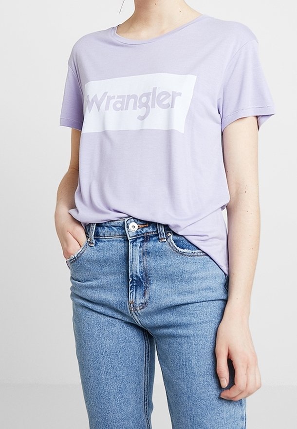 Fialové bavlněné tričko s krátkými rukávy a bílým obd élníkovým grafickým prvkem, na kterém je tučným písmem napsáno "Wrangler". V kombinaci se světle modrými džínami.