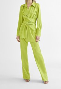 Limegrön, lättviktsomslutande blus med långa ärmar och knytliv, tillsammans med matchande smalbyxor. Slät textur, minimal detaljer.